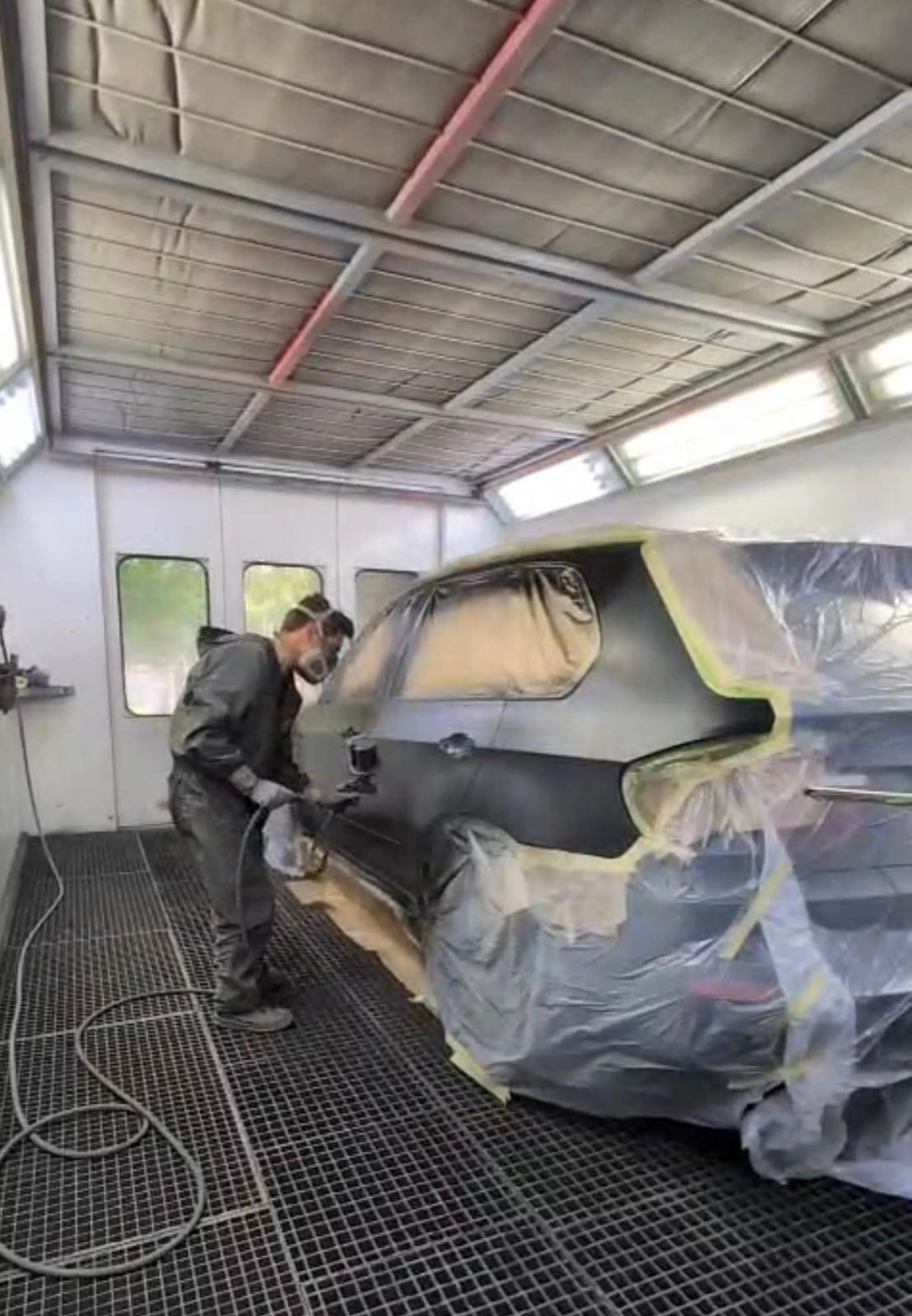 Lackiertechnik John - Fahrzeug Lackierer - Junger hispanischer Mann lackiert in einer Autolackierkabine mit Schutzausrüstung eine Autostoßstange.